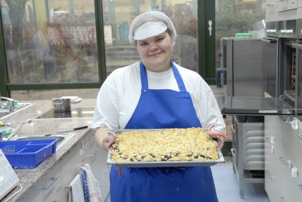 Auszubildende der Hauswirtschaft steht mit einem Blech Kuchen in den Händen in der Küche der Hauswirtschaft